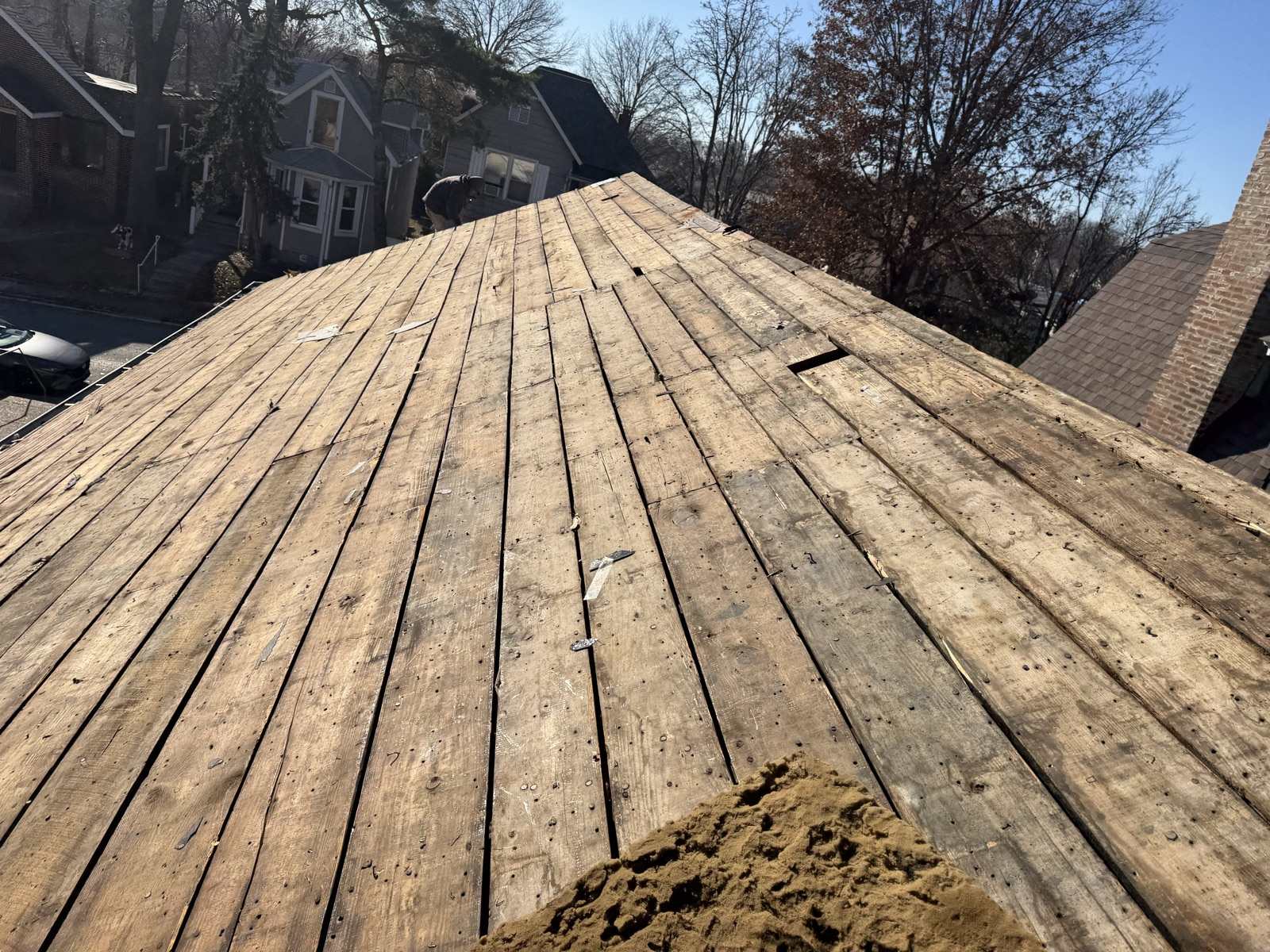 A large roof plane stripped back to bare wooden plank decking studded with old nails, with scraps of felt caught between the boards and a mounded pile of loose tan mineral granules in the foreground, a figure bent over at ground level beyond the roof beside a neighbouring grey house, other houses, a car at the kerb and bare winter trees under a clear blue sky