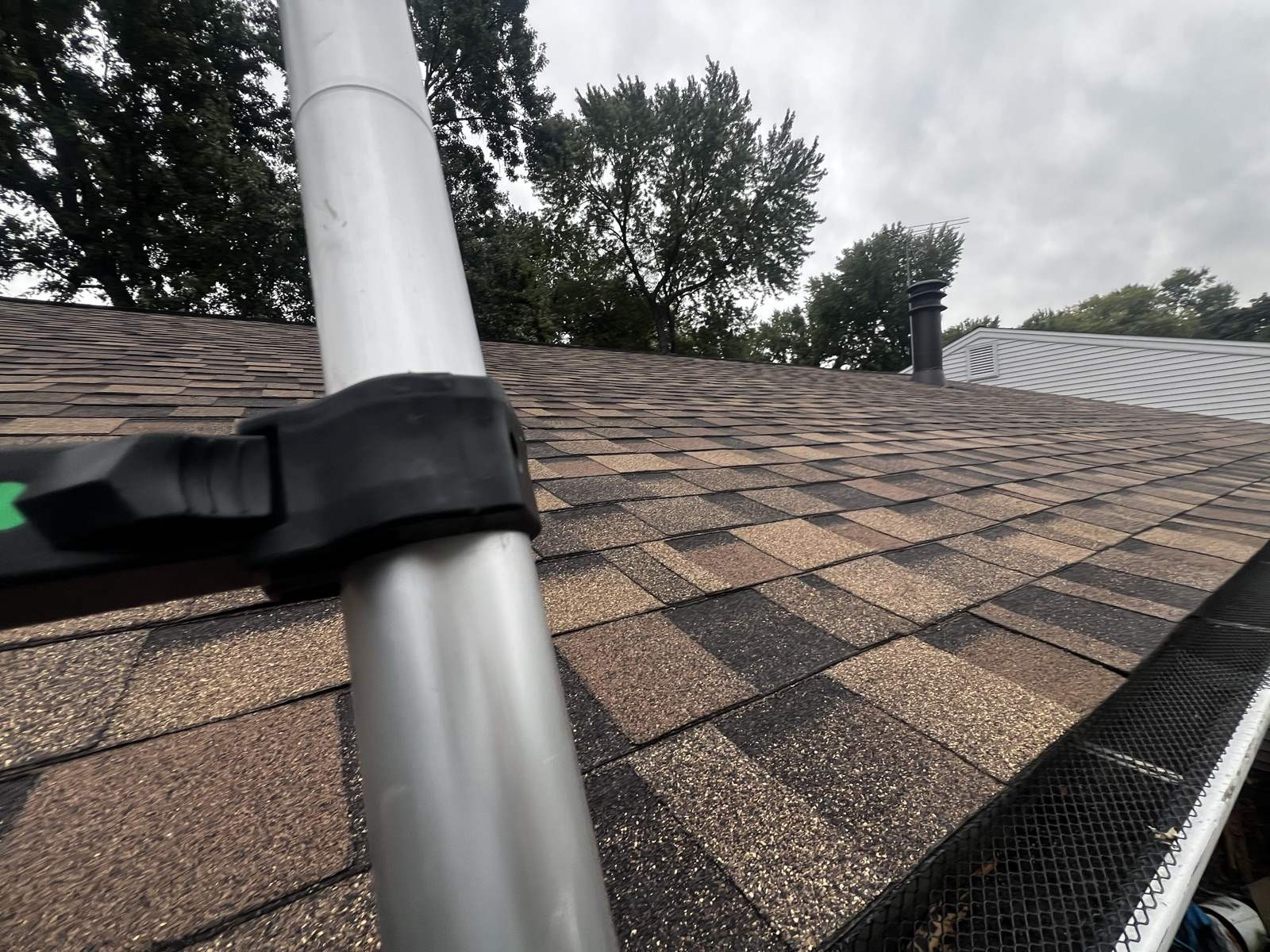 Low view across a weathered brown and tan shingle roof with an aluminium telescoping pole across the foreground and a dark mesh gutter guard along the gutter at the right edge, holding brown leaf litter on its upper surface