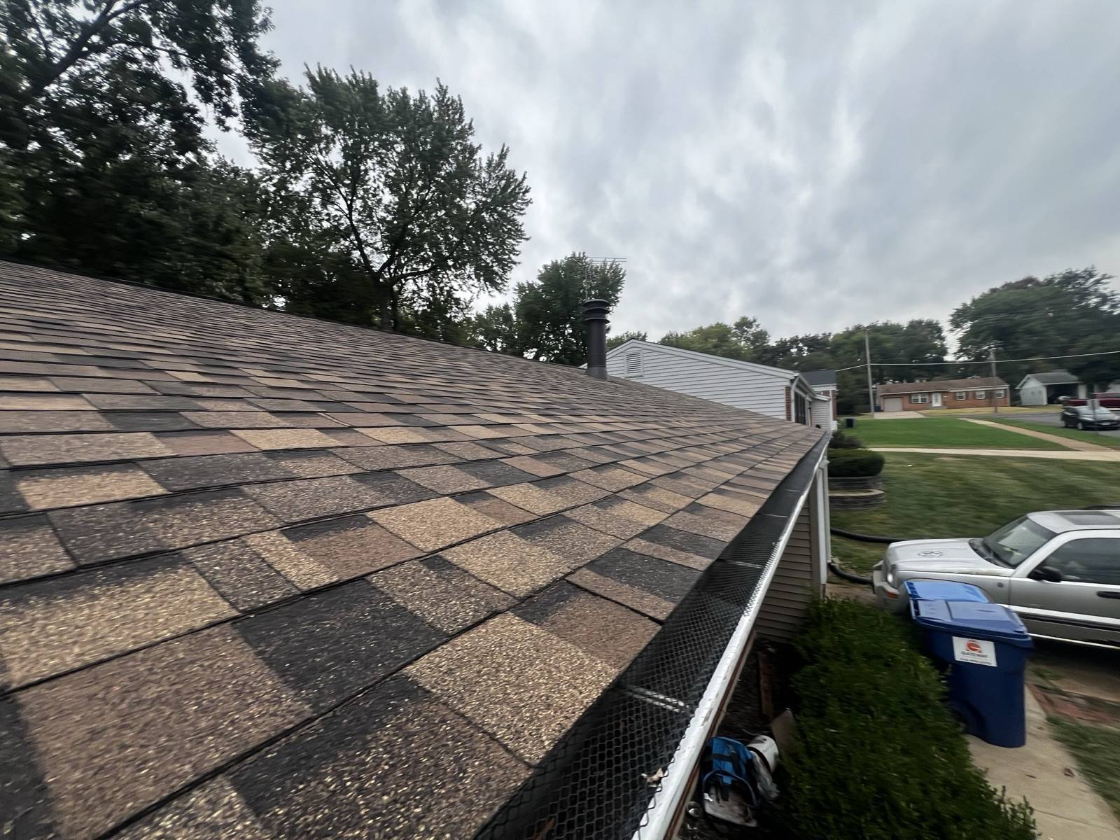 View along the eave of a weathered brown and tan shingle roof on a single storey ranch, with a dark mesh gutter guard fitted along the gutter, a metal flue cap on the ridge, and a driveway with a white pickup, a silver car and a blue wheeled bin below