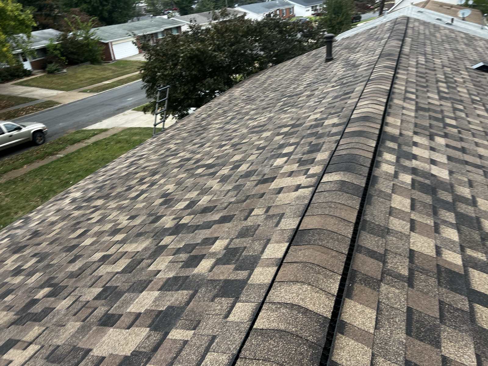 Newly laid brown, tan and charcoal architectural shingle roof photographed along its capped ridge, with a metal flue near the far end, an aluminium ladder standing against the eave at the left, and a suburban street of single storey ranch houses with a white pickup parked at the kerb below