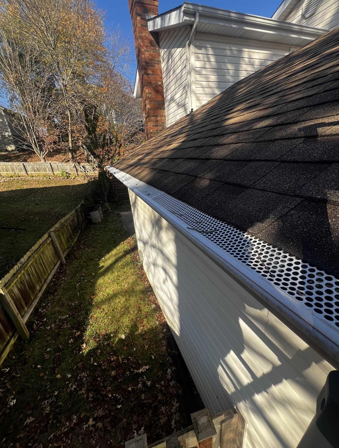 A white perforated metal gutter guard punched with round holes running along the gutter of a dark shingle roof, with two bare twigs and a few small dark scraps of debris lying on top of the perforations, a white sided wall and red brick chimney above, and fallen brown leaves on the lawn below