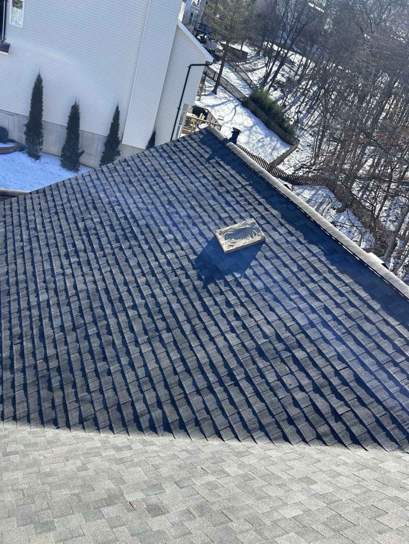 Courses of dark charcoal architectural shingles being laid down a roof plane and meeting a lighter grey shingle surface across the lower part of the frame, with an offcut of new shingle lying loose on the slope, a cream sided neighbouring house, a wooden fence, snow lying on the ground and bare winter trees under hard low sun